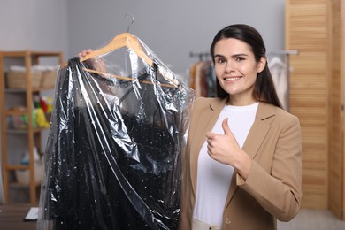 Photo of Dry-cleaning service. Happy woman holding hanger with dress in plastic bag and showing thumb up indoors