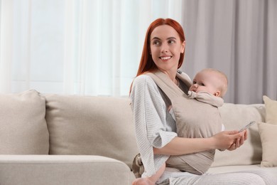 Photo of Mother with smartphone holding her child in sling (baby carrier) at home