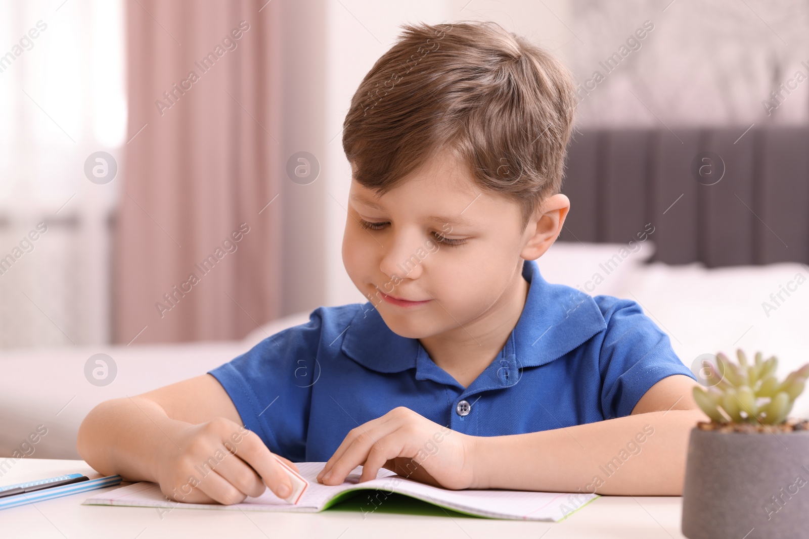 Photo of Little boy erasing mistake in his notebook at white desk indoors