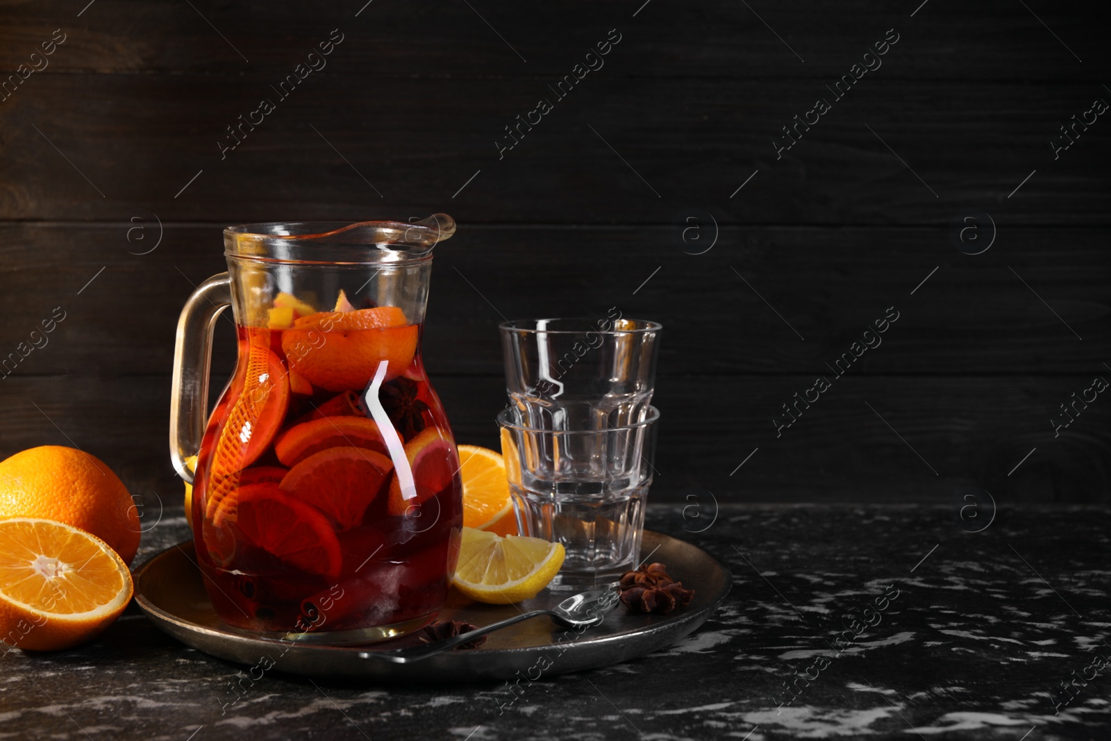 Photo of Glass jug of aromatic punch drink, glasses and ingredients on black table. Space for text