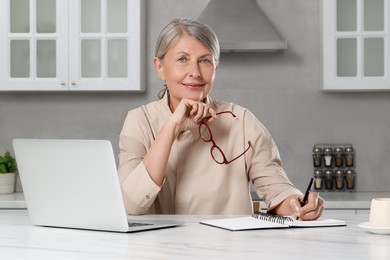 Photo of Beautiful senior woman taking notes near laptop at white marble table in kitchen