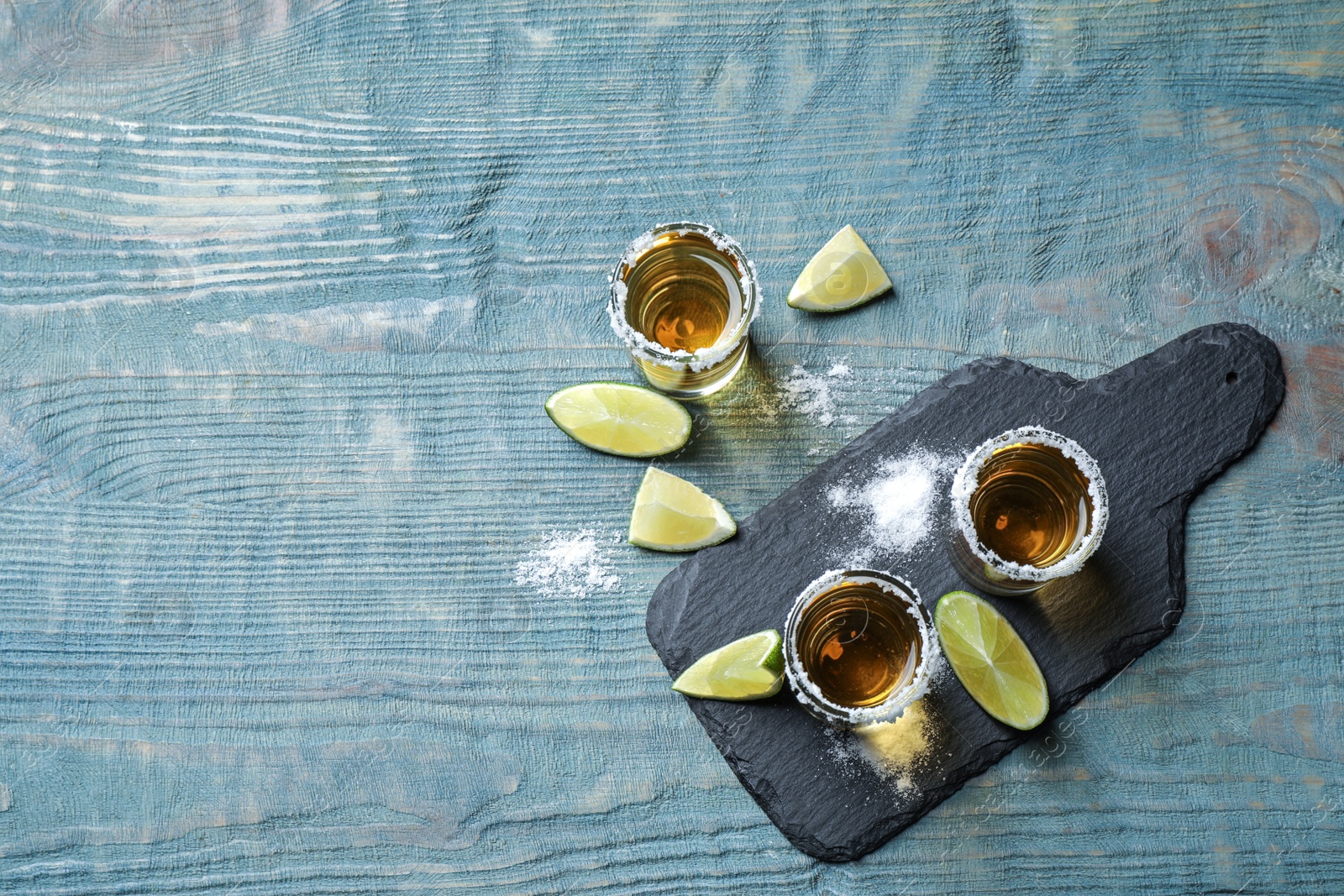Photo of Mexican Tequila shots, lime slices and salt on blue wooden table, flat lay. Space for text