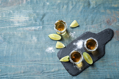 Mexican Tequila shots, lime slices and salt on blue wooden table, flat lay. Space for text