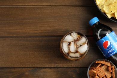 Photo of MYKOLAIV, UKRAINE - FEBRUARY 15, 2021: Glass and bottle of Pepsi with snacks on wooden table, flat lay. Space for text