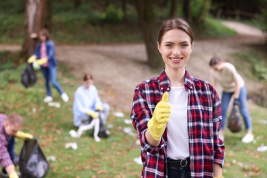 Photo of Young woman in gloves showing thumb up and group of people collecting garbage outdoors