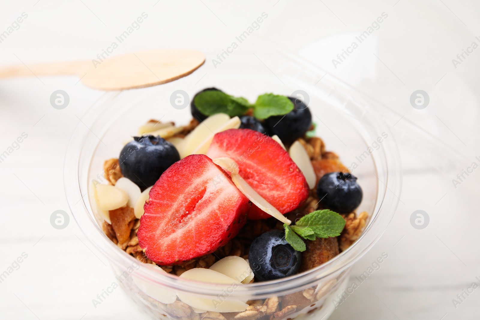 Photo of Tasty granola with berries, almond flakes and mint in plastic cup on white table, closeup