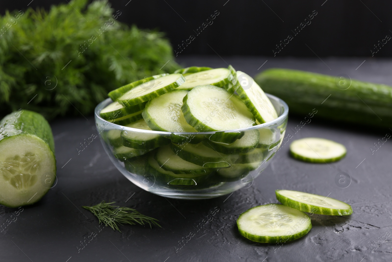 Photo of Cut cucumber in glass bowl on dark gray textured table, closeup