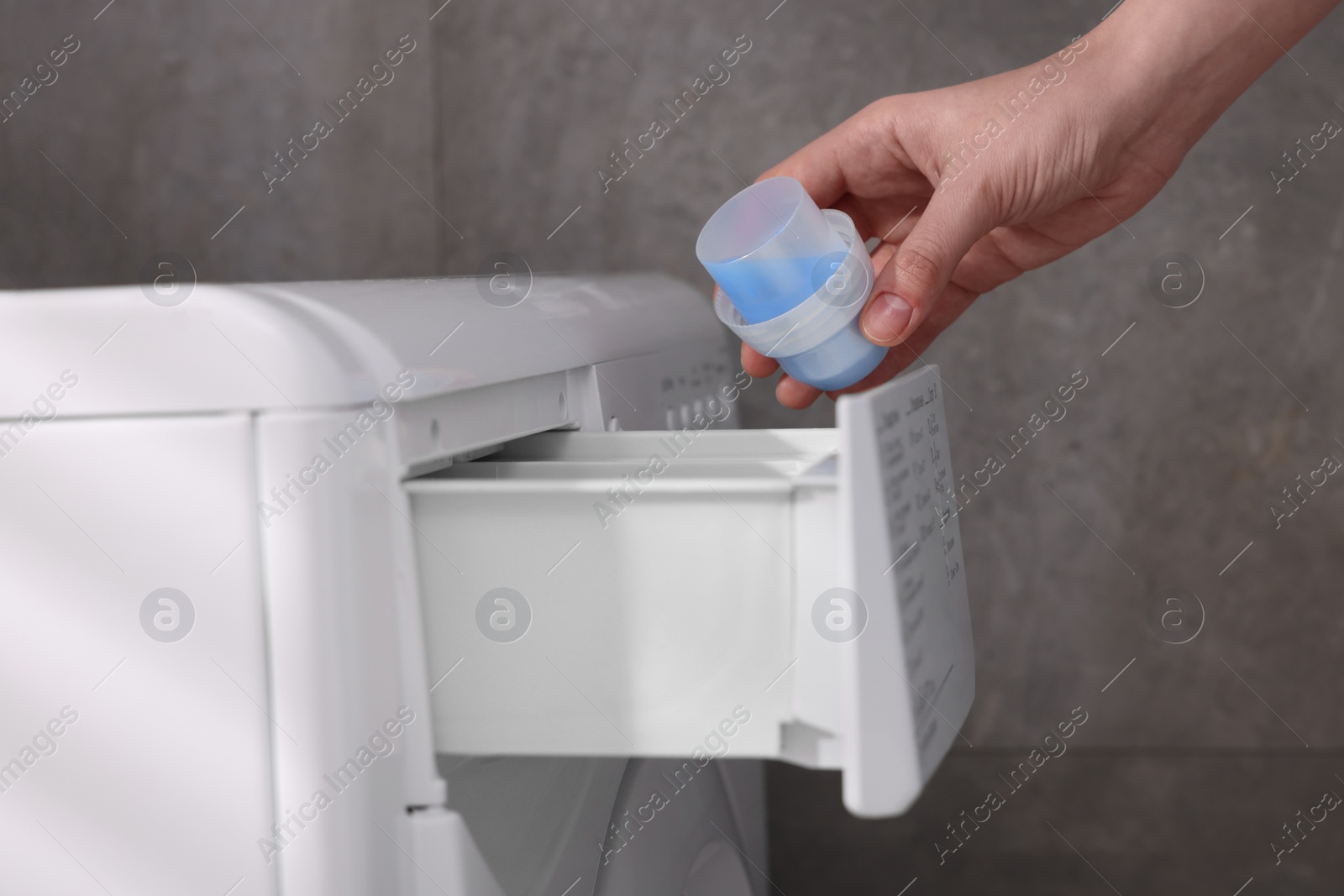 Photo of Woman pouring fabric softener from cap into washing machine near grey wall, closeup