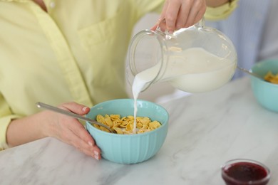 Making breakfast. Woman pouring milk from jug into bowl with cornflakes at white marble table, closeup