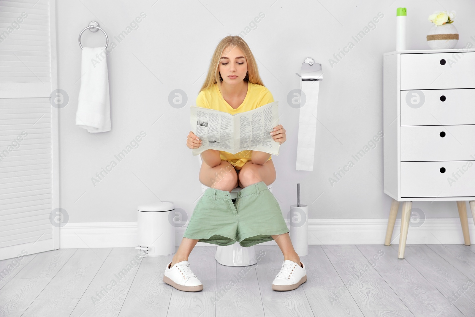 Photo of Young woman reading newspaper while sitting on toilet bowl at home