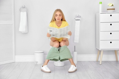 Young woman reading newspaper while sitting on toilet bowl at home
