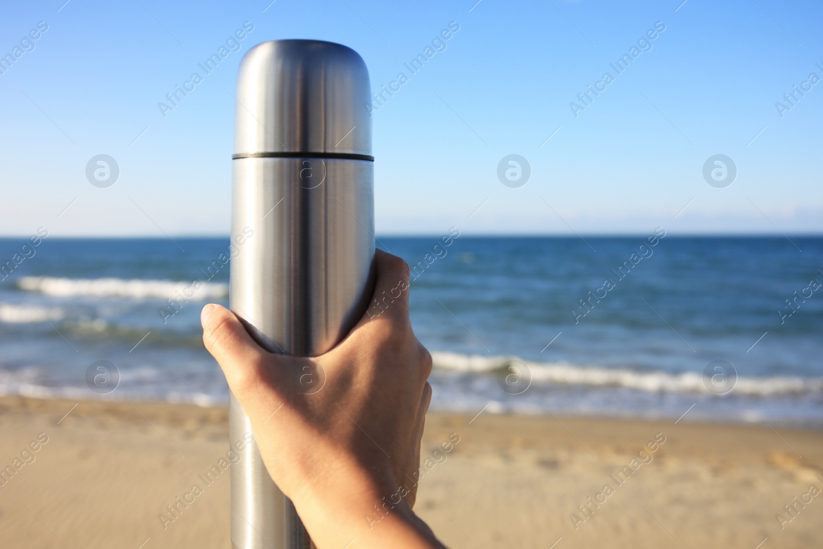 Photo of Woman holding metallic thermos with hot drink on beach near sea, closeup. Space for text