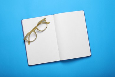 Open notebook and glasses on light blue background, top view