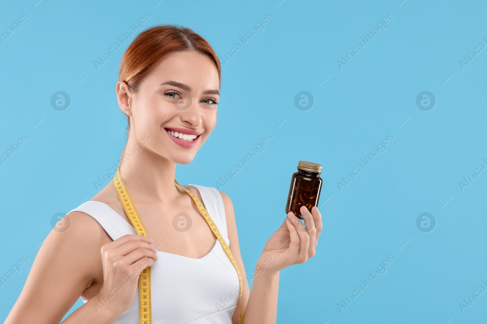 Photo of Happy young woman with bottle of pills and measuring tape on light blue background, space for text. Weight loss