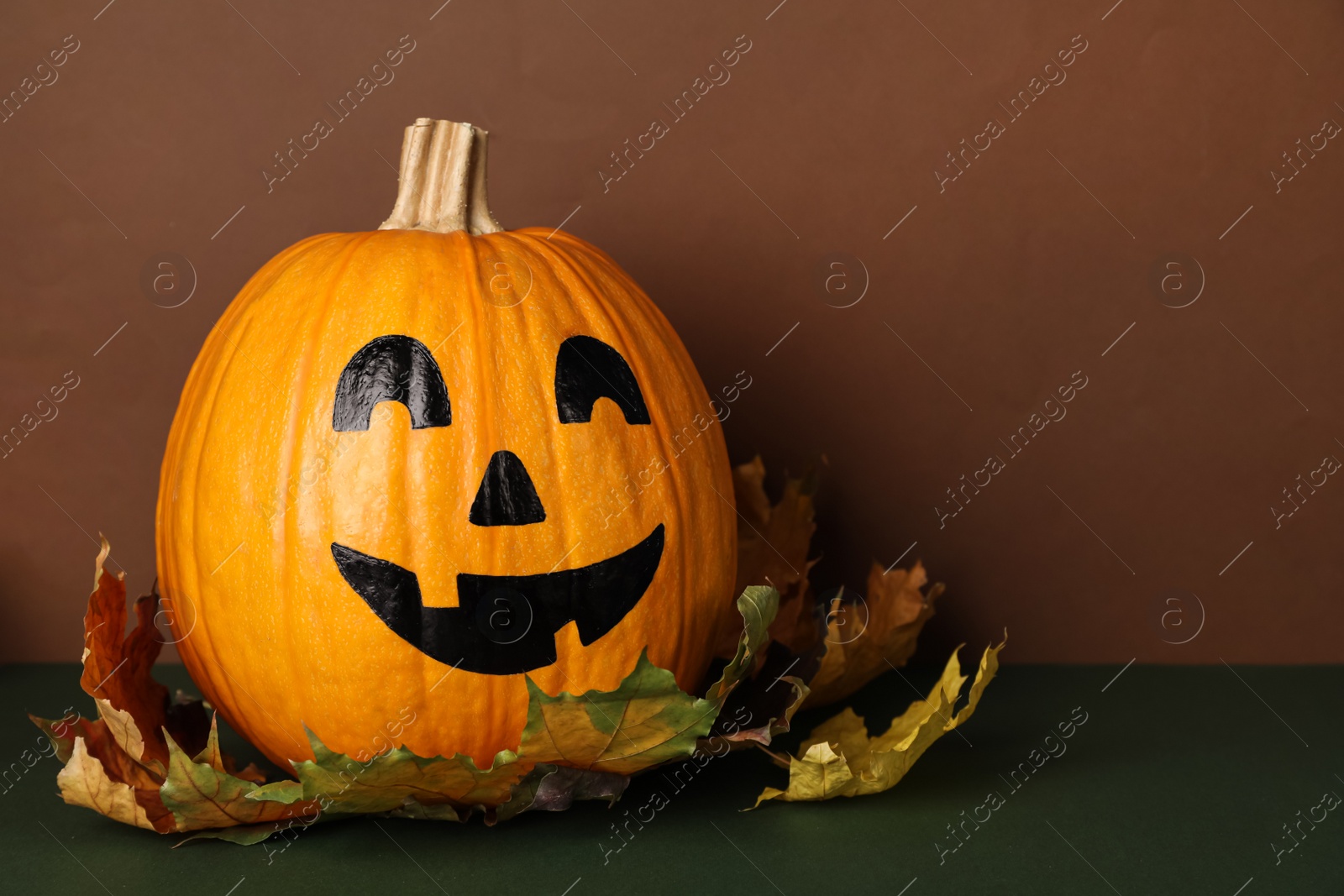 Photo of Pumpkin with drawn spooky face and autumn leaves on color background, space for text. Halloween celebration