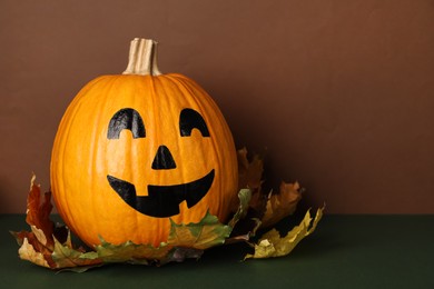 Photo of Pumpkin with drawn spooky face and autumn leaves on color background, space for text. Halloween celebration
