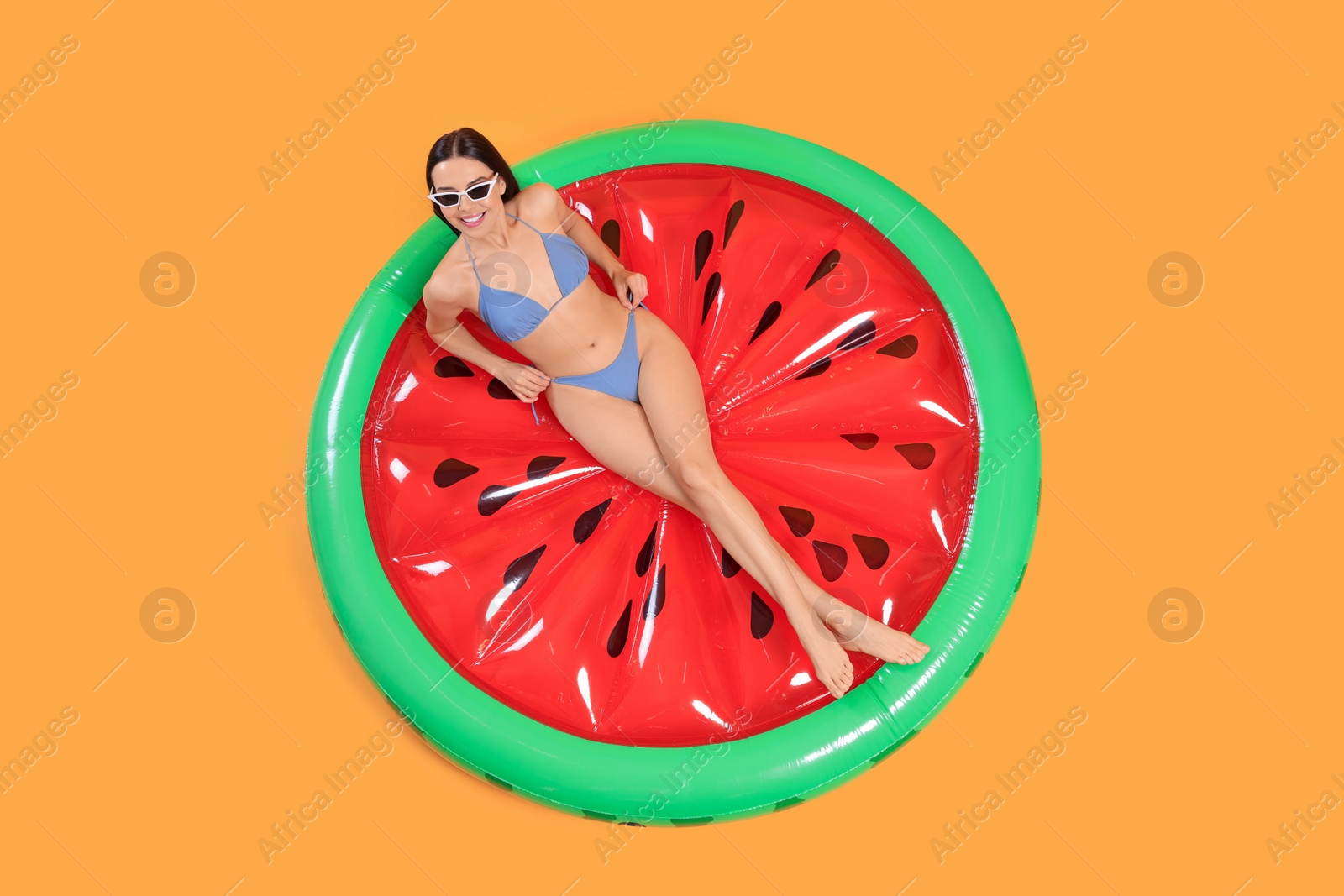 Photo of Happy young woman with beautiful suntan and sunglasses on inflatable mattress against orange background, top view