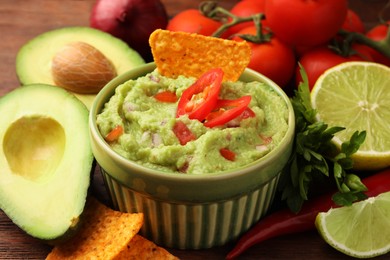 Photo of Bowl of delicious guacamole, nachos chips and ingredients on wooden table, closeup