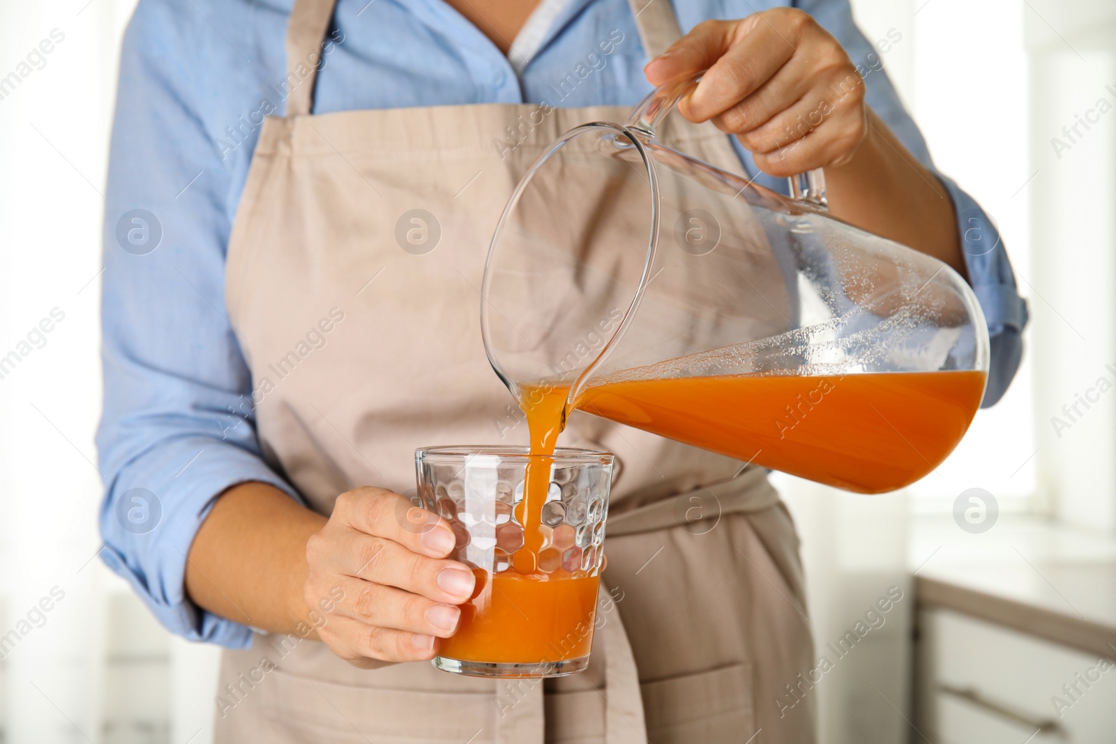 Photo of Woman pouring freshly made carrot juice into glass in kitchen, closeup