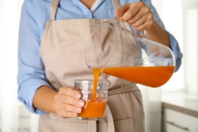 Photo of Woman pouring freshly made carrot juice into glass in kitchen, closeup