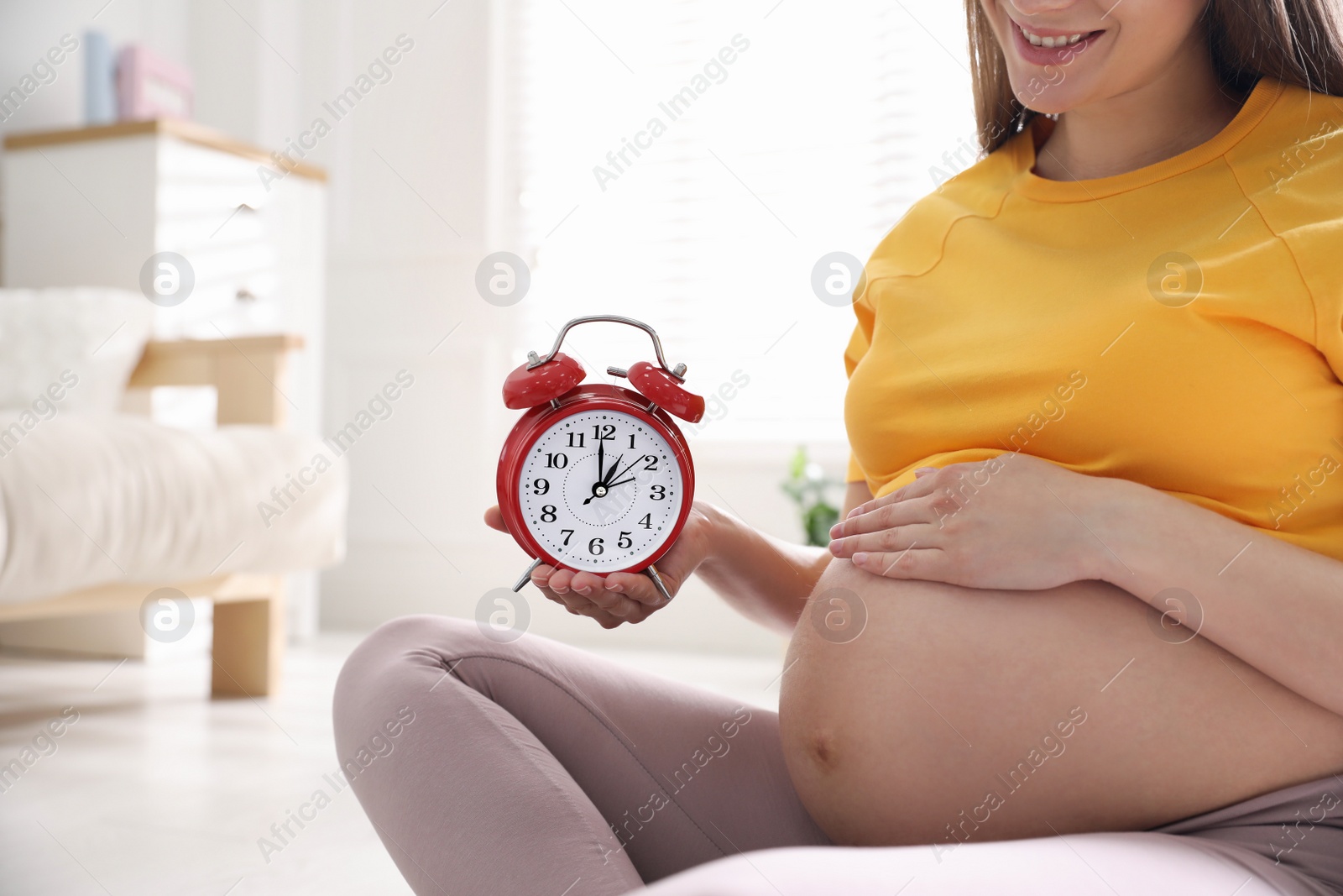 Photo of Young pregnant woman holding alarm clock near her belly at home, closeup. Time to give birth