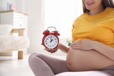 Young pregnant woman holding alarm clock near her belly at home, closeup. Time to give birth