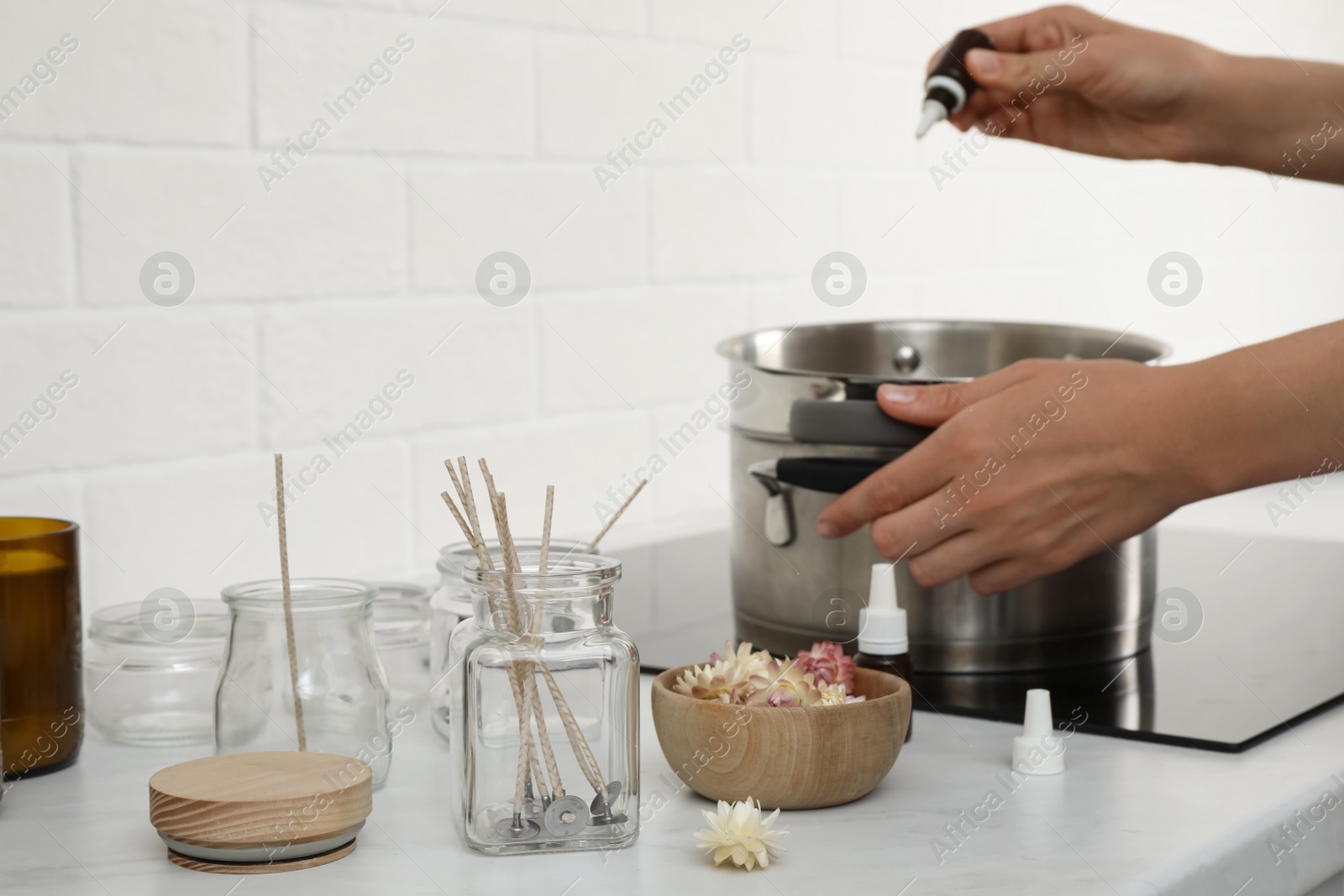 Photo of Woman preparing wax in kitchen, focus on jars with wicks and dry flowers. Making homemade candles