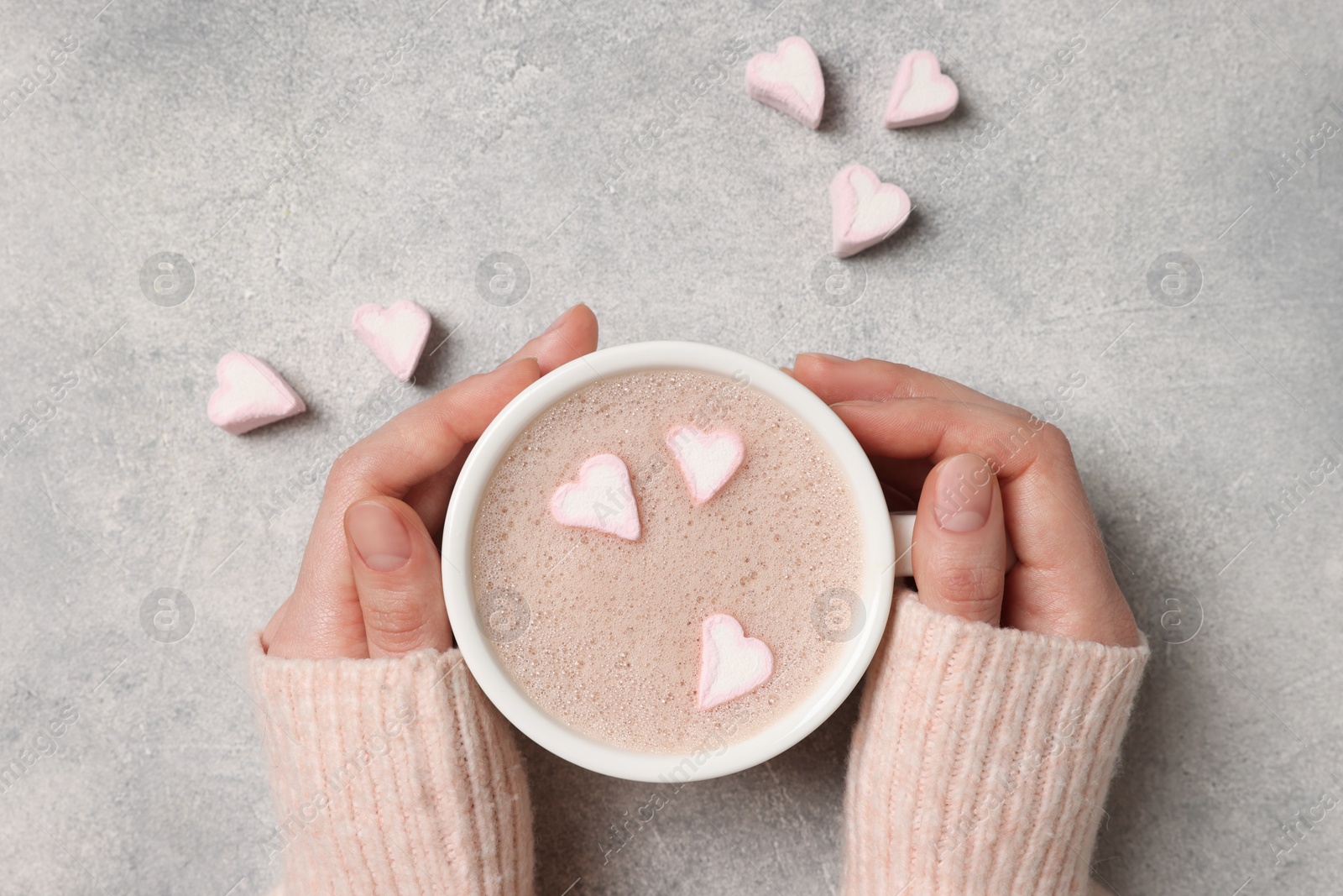 Photo of Woman holding cup of aromatic coffee with heart shaped marshmallows at light grey background, top view