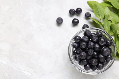 Ripe bilberries in bowl and sprigs with leaves on light marble table, flat lay. Space for text