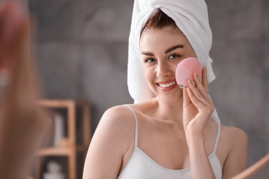 Photo of Washing face. Young woman with cleansing brush near mirror in bathroom