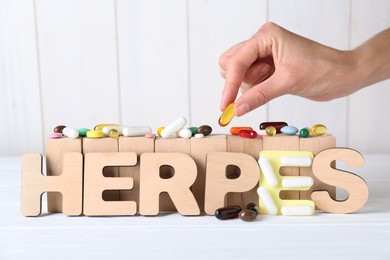 Photo of Woman putting pill near word Herpes on white wooden table, closeup