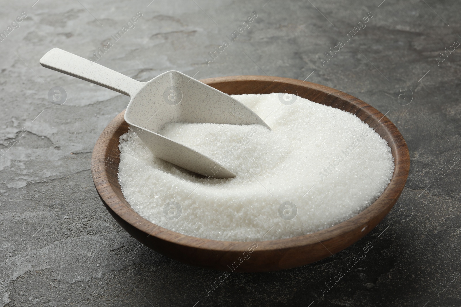 Photo of Granulated sugar in bowl and scoop on grey textured table, closeup