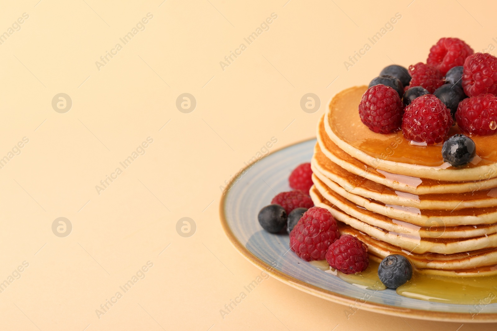 Photo of Stack of tasty pancakes with raspberries, blueberries and honey on pale orange background, closeup. Space for text