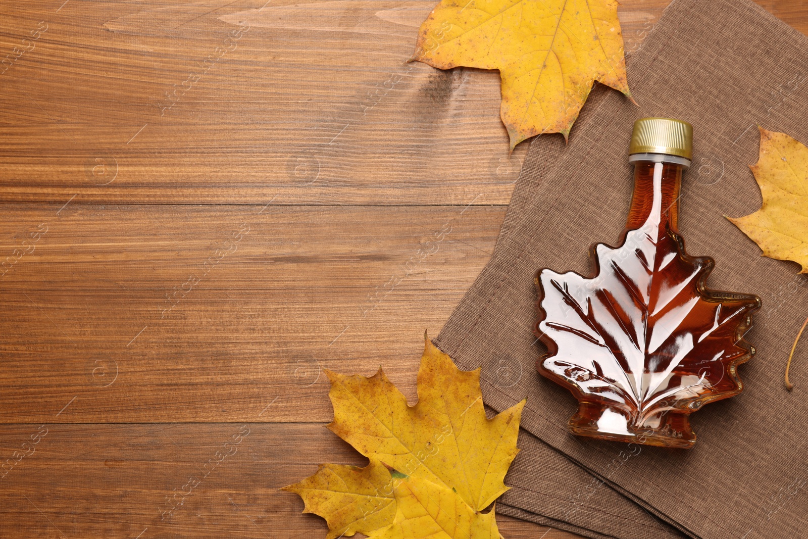 Photo of Bottle of tasty maple syrup and dry leaves on wooden table, flat lay. Space for text