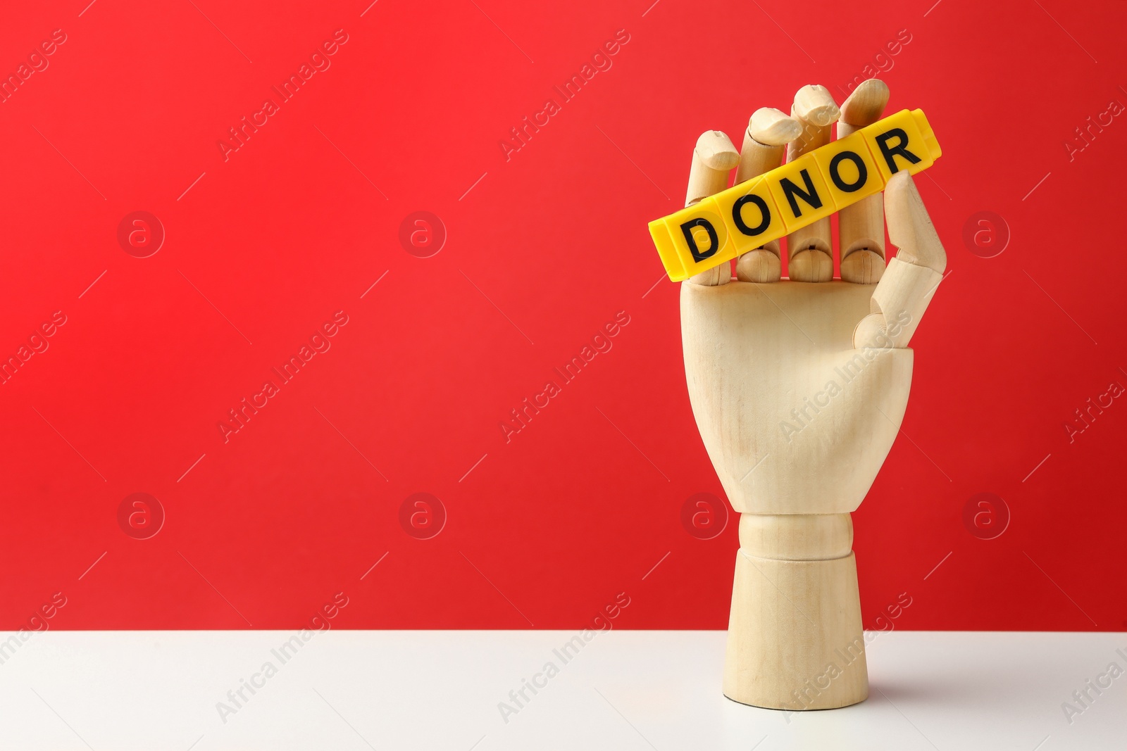 Photo of Mannequin hand holding word Donor made of cubes on white table against red background. Space for text