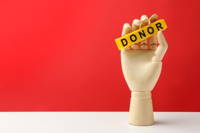 Photo of Mannequin hand holding word Donor made of cubes on white table against red background. Space for text