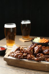 Photo of Tasty chicken wings, ketchup, glasses of beer and vegetable sticks on wooden table. Delicious snacks