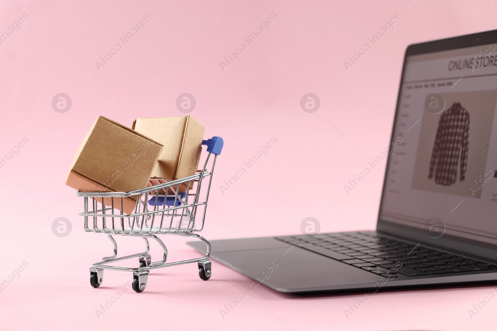 Photo of Online store. Laptop, mini shopping cart and parcels on pink background