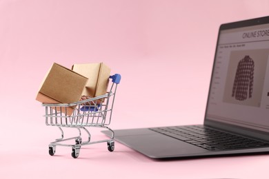 Photo of Online store. Laptop, mini shopping cart and parcels on pink background
