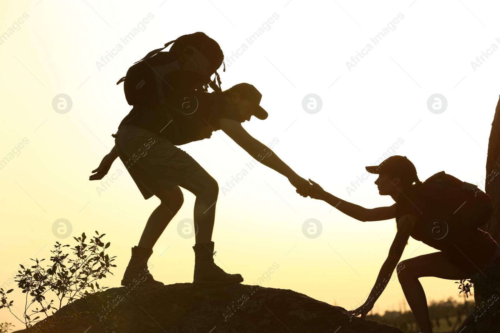 Photo of Silhouettes of man and woman helping each other to climb on hill against sunset