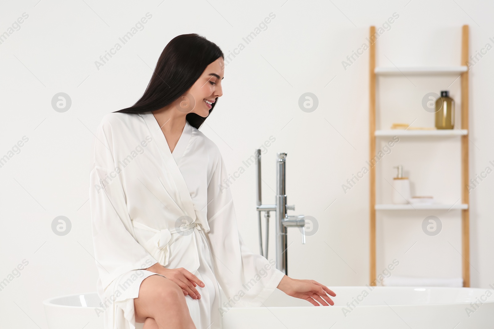 Photo of Beautiful happy woman in stylish bathrobe sitting on tub in bathroom