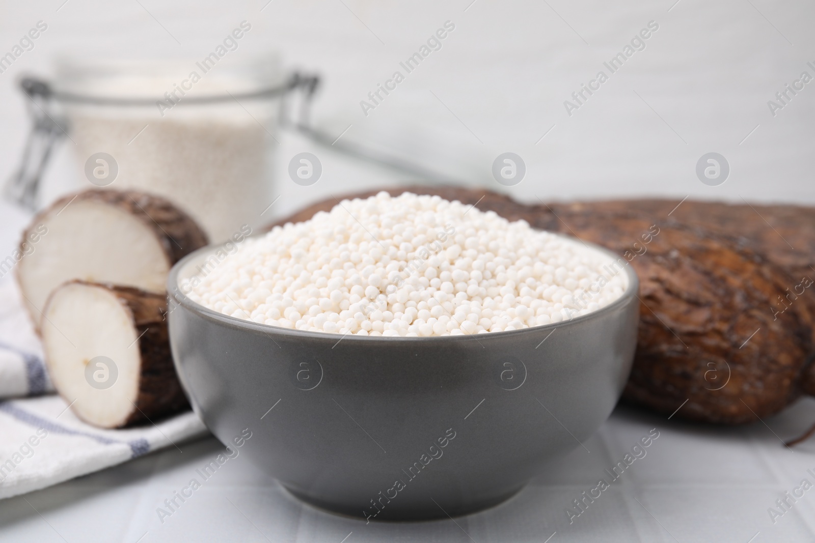 Photo of Tapioca pearls in bowl and cassava roots on white tiled table, closeup