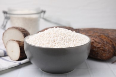 Tapioca pearls in bowl and cassava roots on white tiled table, closeup