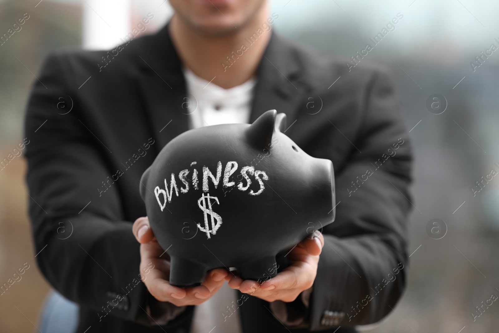 Photo of Man holding piggy bank with word BUSINESS against blurred background, closeup