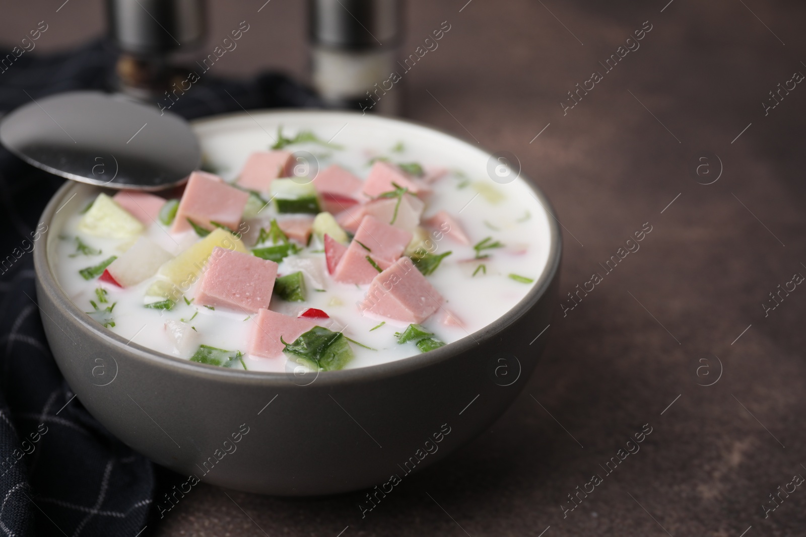 Photo of Delicious cold summer soup (okroshka) with boiled sausage in bowl and spoon on brown table, closeup. Space for text