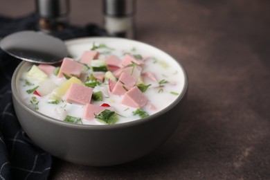 Delicious cold summer soup (okroshka) with boiled sausage in bowl and spoon on brown table, closeup. Space for text