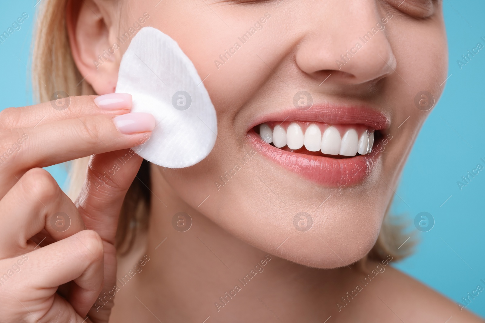 Photo of Young woman cleaning face with cotton pad on light blue background, closeup