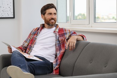 Photo of Smiling handsome bearded man with magazine looking away on sofa indoors