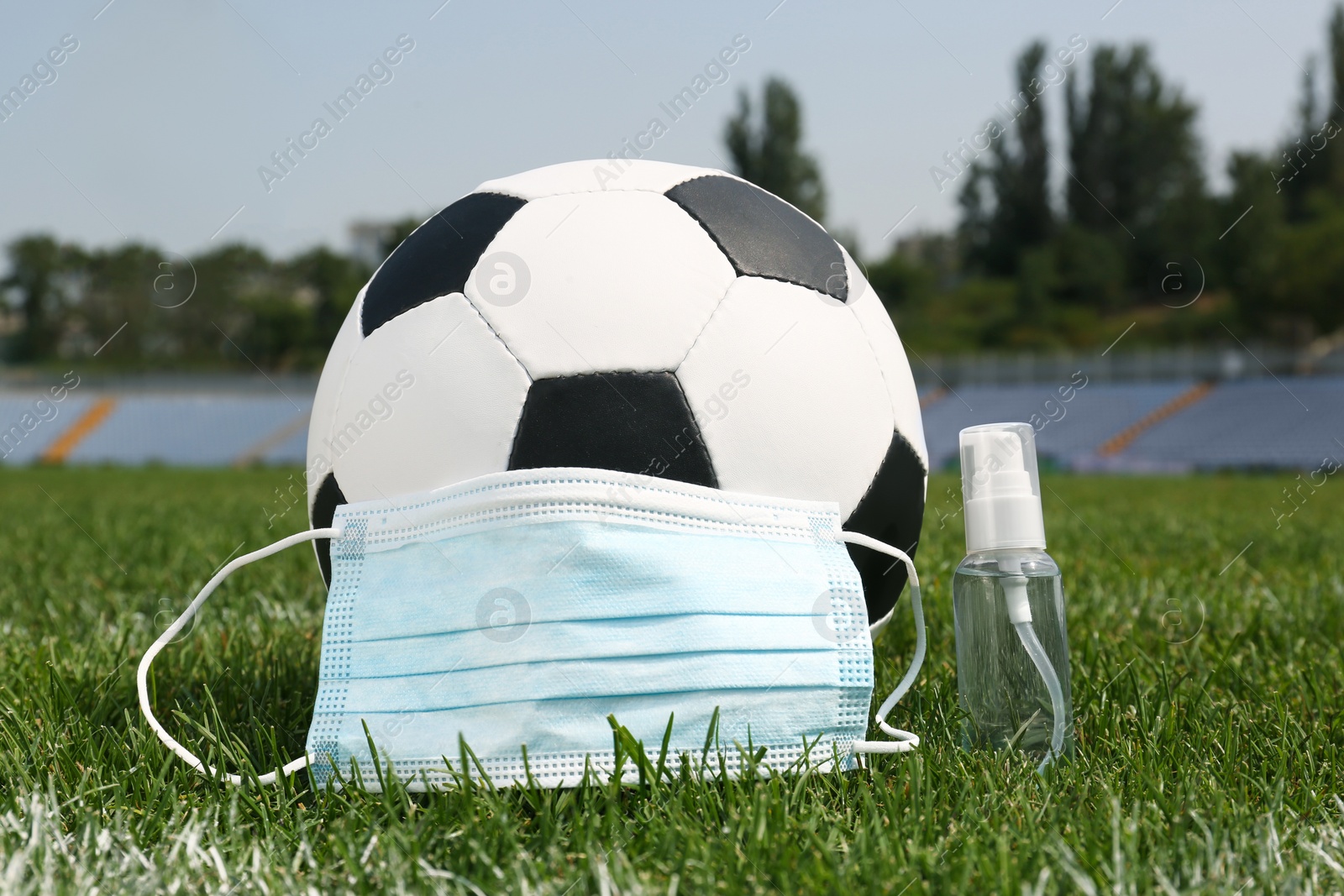 Photo of Football ball with hand sanitizer and protective mask on green field grass in stadium