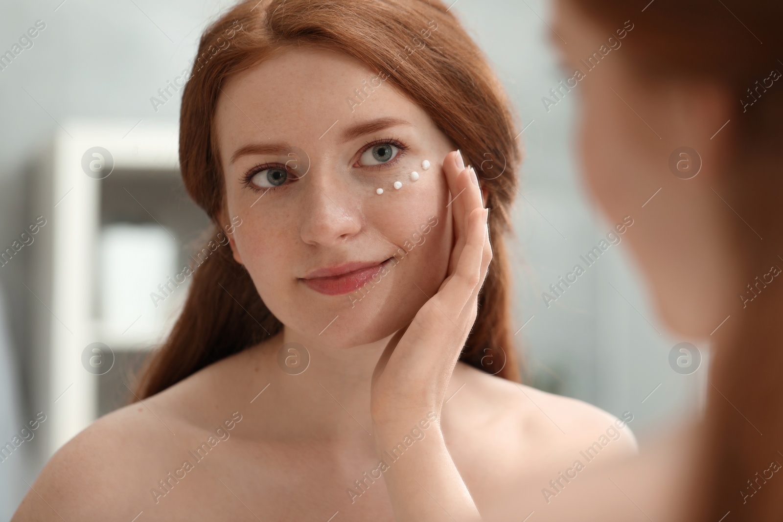 Photo of Beautiful woman with freckles applying cream onto her face near mirror in bathroom